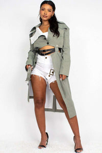 A model is posing in a fashion-forward outfit, consisting of a sage green trench coat worn off the shoulder over a white cropped tank top and distressed white denim shorts. The coat features traditional details like a collar, lapels, and cuffed sleeves with button accents. She accessorizes with a black belt featuring multiple silver buckles. Her hair is styled in loose waves, and her makeup is natural, complementing the casual yet chic vibe of the outfit.