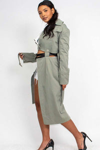 A model is posing in a fashion-forward outfit, consisting of a sage green trench coat worn off the shoulder over a white cropped tank top and distressed white denim shorts. The coat features traditional details like a collar, lapels, and cuffed sleeves with button accents. She accessorizes with a black belt featuring multiple silver buckles. Her hair is styled in loose waves, and her makeup is natural, complementing the casual yet chic vibe of the outfit.