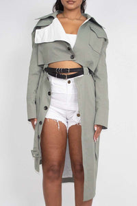 A model is posing in a fashion-forward outfit, consisting of a sage green trench coat worn off the shoulder over a white cropped tank top and distressed white denim shorts. The coat features traditional details like a collar, lapels, and cuffed sleeves with button accents. She accessorizes with a black belt featuring multiple silver buckles. Her hair is styled in loose waves, and her makeup is natural, complementing the casual yet chic vibe of the outfit.