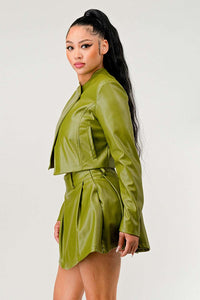 olive green faux leather jacket and mini skirt. The jacket has a unique asymmetrical cut with a collarless design and a single button near the shoulder. The high-waisted skirt flares slightly at the hem, and a thin belt accentuates the waist. She completes the look with a simple white crop top and clear strap heeled sandals, presenting a chic and modern appearance.