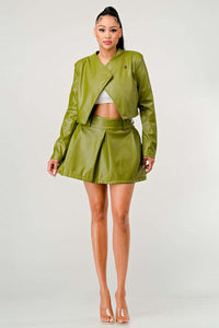 olive green faux leather jacket and mini skirt. The jacket has a unique asymmetrical cut with a collarless design and a single button near the shoulder. The high-waisted skirt flares slightly at the hem, and a thin belt accentuates the waist. She completes the look with a simple white crop top and clear strap heeled sandals, presenting a chic and modern appearance.