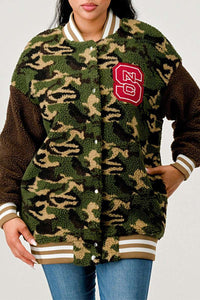 Fuzzy knit camouflage cardigan with ribbed cuffs and a hem in shades of green and brown. The cardigan is adorned with a large red and white varsity-style letter patch on the left side of the chest. It's paired with a collared shirt and denim jeans, suggesting a casual yet trendy look.