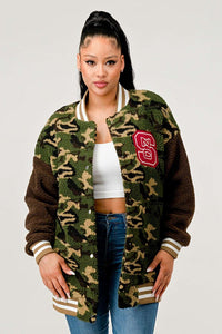 Fuzzy knit camouflage cardigan with ribbed cuffs and a hem in shades of green and brown. The cardigan is adorned with a large red and white varsity-style letter patch on the left side of the chest. It's paired with a collared shirt and denim jeans, suggesting a casual yet trendy look.