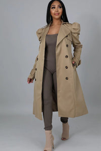 A woman stands confidently, modeling a neutral-colored classic trench coat that is open to reveal a coordinating taupe jumpsuit beneath. The coat features a double-breasted design with large buttons, epaulettes, and a belt. She completes the ensemble with open-toe heeled ankle boots in a matching taupe shade.